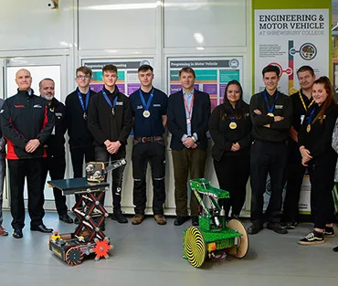 A group of apprentices, College staff, including the Principal and employers standing behind two robots